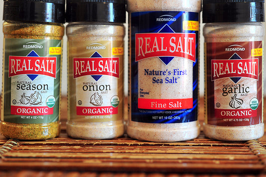 Four shakers of Real Salt brand seasoning salts, labeled as Organic Garlic, Organic Onion, Fine Salt, and Seasoning Salt, displayed on a surface with the brand name 'REAL SALT' prominently visible.