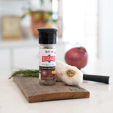 A shaker bottle of Redmond Real Salt Organic Garlic Pepper on a wooden board with a piece of garlic and green spices in the background.